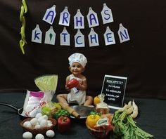 a baby doll sitting on top of a table surrounded by fruits and vegetables in front of a sign that says arrhas kitchen