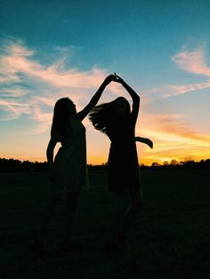 two girls are standing in the grass with their arms up as the sun sets behind them