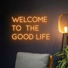 a neon sign that reads welcome to the good life next to a chair and plant