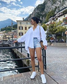 a woman standing next to a body of water with buildings in the background and mountains behind her