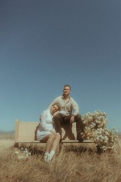 a man and woman sitting on a bench in the middle of a field with wildflowers