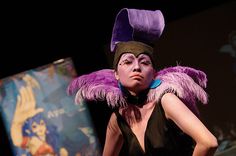 a woman with purple makeup and feathers on her head is standing in front of a poster