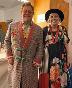 two people standing next to each other in front of a wooden paneled wall wearing colorful clothing and hats