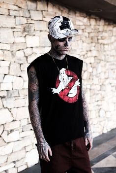 a man with tattoos wearing a black shirt and hat standing in front of a stone wall