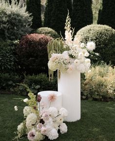 a tall white vase filled with flowers on top of a lush green field next to shrubbery