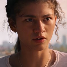 a close up of a person wearing a necklace and looking off into the distance with buildings in the background