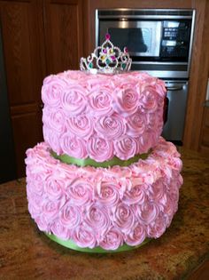 two tiered cake with pink frosting roses and a crown on top, sitting on a kitchen counter