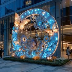 people walk past a large illuminated sculpture in the city at night, decorated with flowers and butterflies