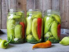 three jars filled with pickles and peppers on top of a table next to green peppers