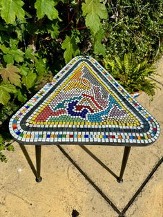 a mosaic table sitting on top of a cement ground next to green plants and trees