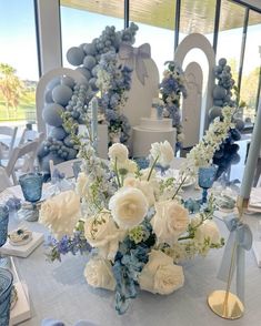 a centerpiece with white and blue flowers on a table in front of large windows