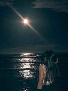 a woman standing in the ocean at night with her back turned to the camera, looking up at the moon