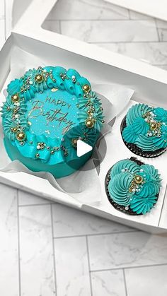 two cupcakes with blue frosting and gold decorations in a white box on the floor