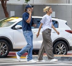 two people walking across the street while talking on their cell phones and one is holding a phone up to his ear