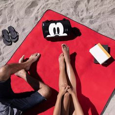 two people laying on a red towel at the beach with their feet up in the sand