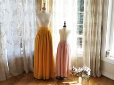 two dresses on mannequins in front of window with curtains and flowers next to them