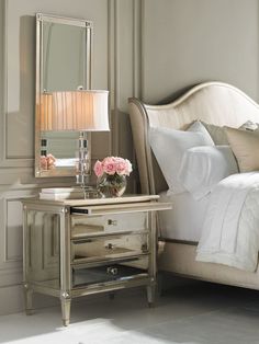 a white bed sitting next to a mirror and lamp on top of a wooden table