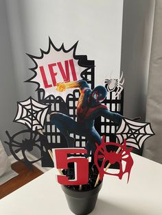 a spiderman cake topper sitting in a flower pot on a table next to the letter d