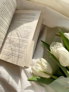 an open book sitting on top of a bed next to white flowers