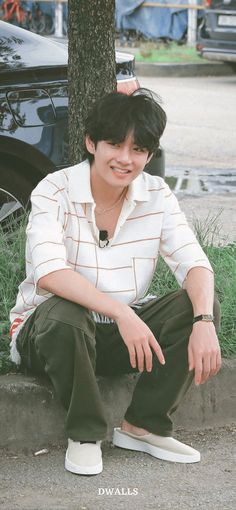 a young man sitting on the ground in front of a tree with his hand on his hip