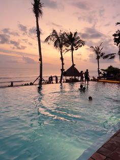 people are playing in the swimming pool at sunset or dawn with palm trees and thatched umbrellas