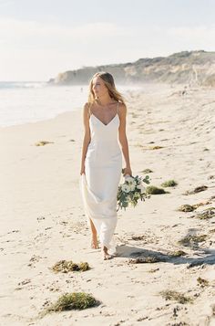 a woman is walking on the beach with flowers in her hand and wearing a white dress