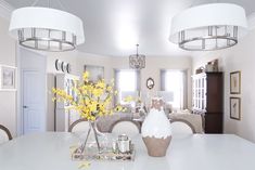 a white dining room table with yellow flowers in vases on it and two chandeliers hanging from the ceiling