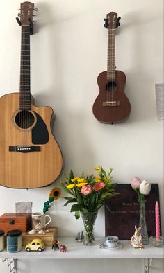 two guitars are hanging on the wall above a table with flowers in vases and other items