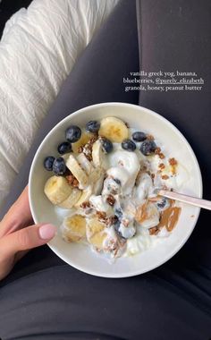 a person holding a bowl of cereal with bananas and blueberries in it while sitting on a seat