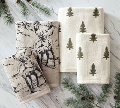 three hand towels with moose and pine trees printed on them next to evergreen cones, firs and pine needles