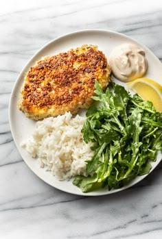 a white plate topped with meat, rice and greens next to a side of lemon wedges