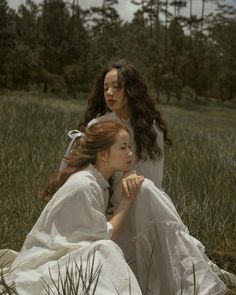 two women in white dresses sitting on the ground next to each other with trees in the background