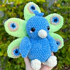 a hand holding a blue and green crocheted stuffed animal in front of some bushes