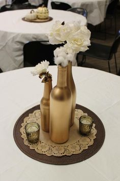 three gold vases with white flowers in them on a doily at a table