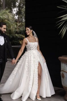 a bride and groom holding hands in front of a black wall wearing tuxedos