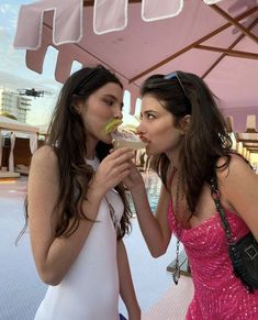 two women standing next to each other in front of a pink umbrella eating food from plates