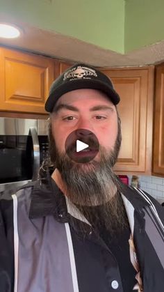 a man with a long beard wearing a baseball cap and jacket in a kitchen next to wooden cabinets