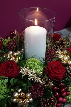 a white candle surrounded by red roses and greenery in a glass vase on a table