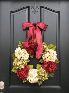 a wreath with red and white flowers is hanging on a black front door that has a pink ribbon