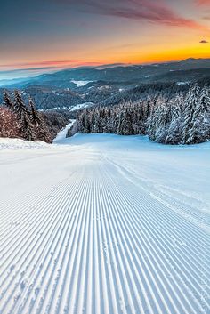 the sun is setting over a snowy mountain with tracks in the snow and trees on both sides