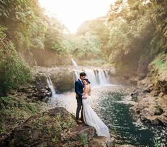 a bride and groom standing on the edge of a waterfall with their arms around each other