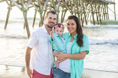 a man and woman holding a baby standing on the beach with a pier in the background