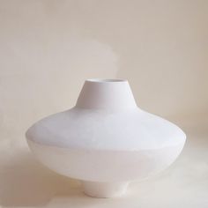 a white vase sitting on top of a table next to a light colored wall behind it