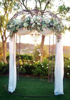 an outdoor wedding ceremony setup with white drapes and flowers on the arch, surrounded by greenery