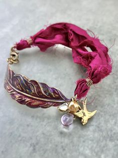 WHAT IT IS: Handmade bohemian-style bracelet featuring upcycled magenta sari silk, a large hand-enameled and hand-formed brass feather, a brass bird charm, a small vintage Swarovski channel-set crystal, and vintage beads. THE SPECS: WHY IT'S SPECIAL: The brass feather used in this bracelet was enameled by hand in shades of magenta, purple, burgundy, and blue. The ribbon is upcycled from torn and dyed silk that was leftover from the sari manufacturing process. Purple Bracelets For Festivals And Gifts, Sari Ribbon Jewelry, Silk Ribbon Jewelry, Sari Silk Jewelry, Brass Bird, Sari Ribbon, Artisan Jewelry Handmade, Purple Burgundy, Silk Jewelry