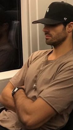 a man sitting on a train with his arms crossed and wearing a baseball cap, looking out the window