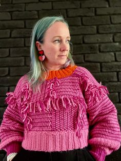 a woman with blue hair wearing a pink sweater and black skirt, standing in front of a brick wall