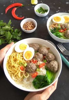 someone holding a bowl of noodles with meatballs and veggies in it on a table