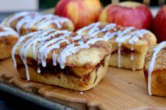 cinnamon roll with icing sitting on top of a wooden cutting board next to apples