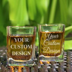 two shot glasses with the words your custom design on them sitting on a table in front of some plants